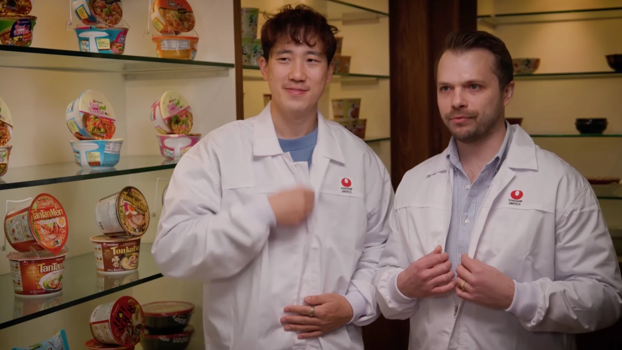 Two hosts in Nongshim America lab coats standing in front of shelves of Nongshim bowl noodles