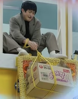 A scene from the series: a cast member on a window ledge tying up a custom Nongshim box with yellow rope